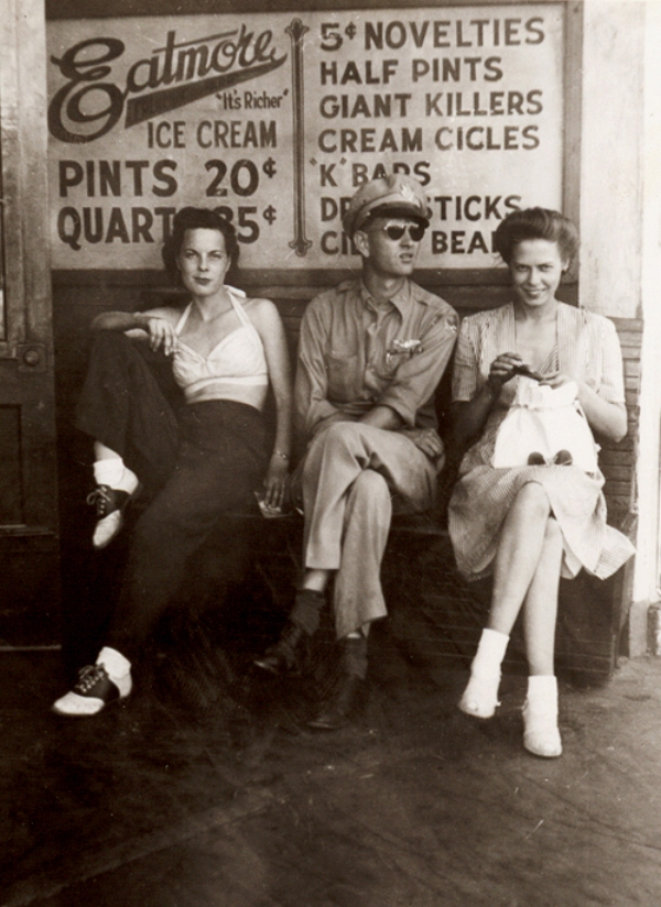 1940s vintage photo of a woman wearing pants, swimsuit top and saddle shoes sitting next to a man in uniform and another woman in 1940s dress. 