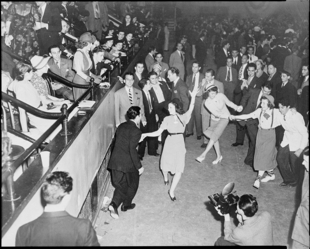 1940s vintage photo of a group of people dancing the Lindy hop at a show. They are wearing 1940s fashions. 