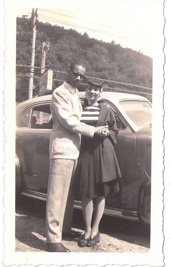 1940s vintage photo of a stylish Black Man and Black Woman posing in front of their car in 1940s fashions.