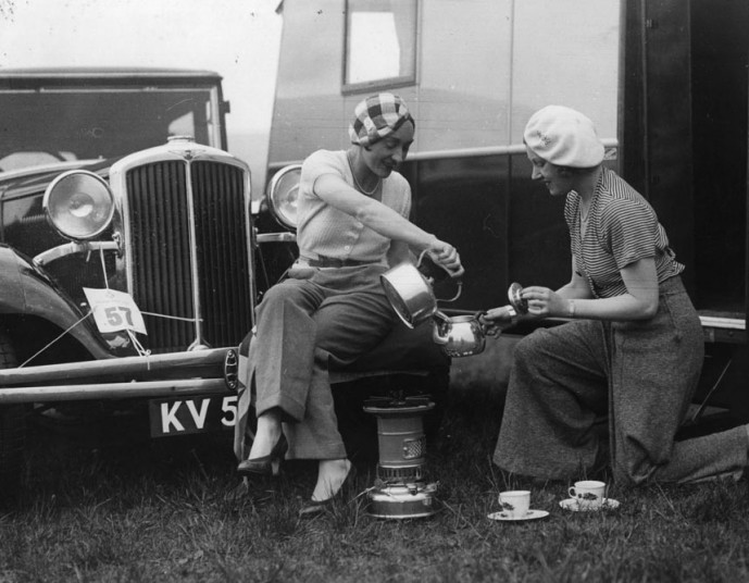 1930's vintage photo of 2 women in 1931 in berets and 1930s fashions making tea outside of their camper in front of a 1930s vehicle.