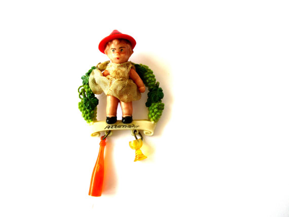 The hatpin is from a wine region near the Rhine River; Altenahr is situated between Bonn and Koblenz. The little girl doll is standing against a background of grapes, and a yellow wine glass and orange wine bottle dangle beneath the main portion of the pin. This was likely a souvenir from a 1950s era European vacation.