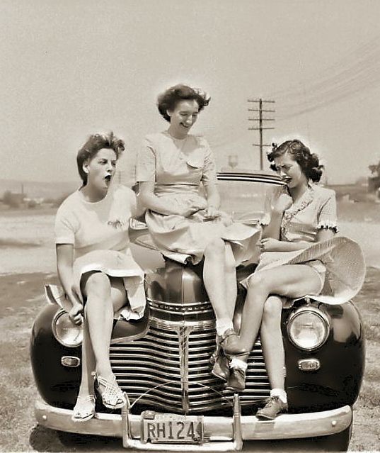 1940s vintage image of 3 woman getting ready to pose on a car but then a gust of wind hits them. 