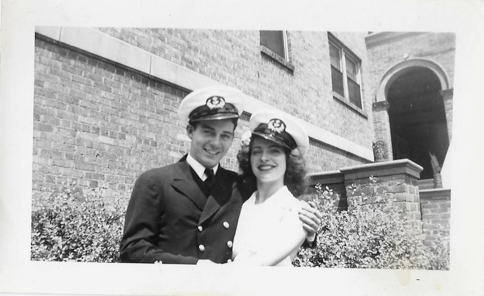 1940s vintage photo of a young man and woman holding each other with Navy hats on their heads during WW2. The young woman is wearing a hair flower in her hair. 