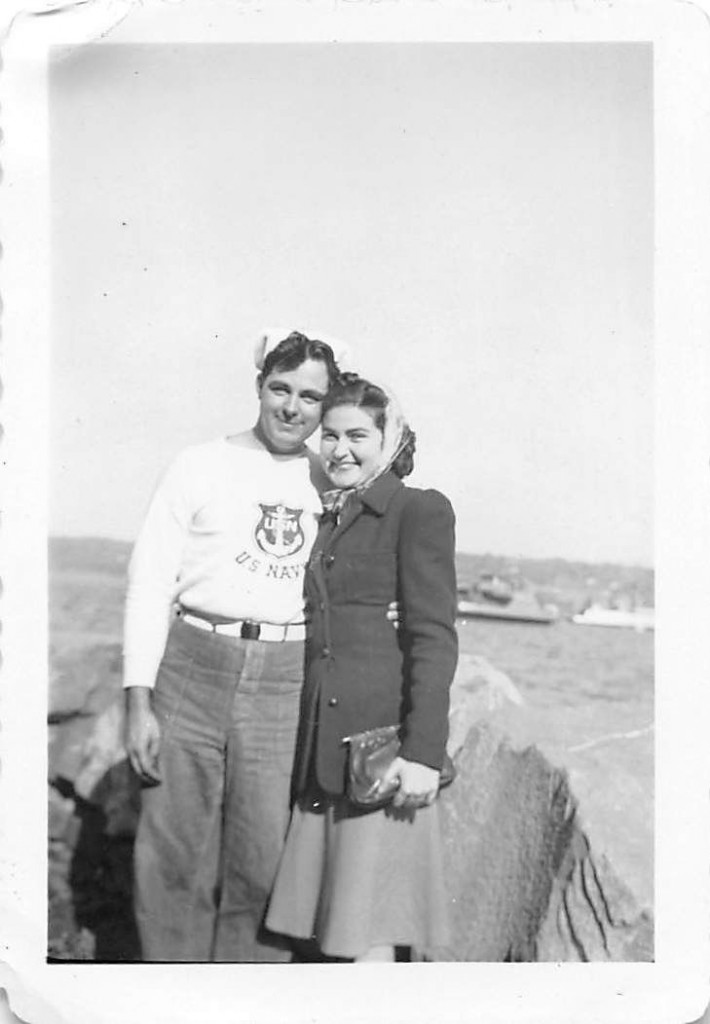 1940s vintage photo of a young couple posing together. He is in a US navy sweater and navy hat and the young woman is in a jacket and hair scarf. 