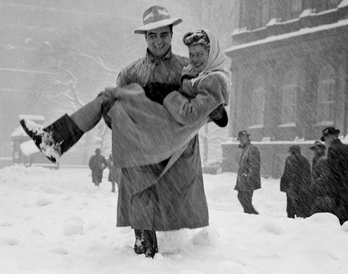 1947 photo in New York city of a man carrying a woman thru the snowstrom. Both are wearing 1940s winter fashions.