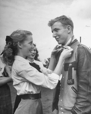 1940s vintage photo of a young woman in 1946 tieing a scarf around a young man wearing a varsity jacket. 1940s Fashion inspiration.