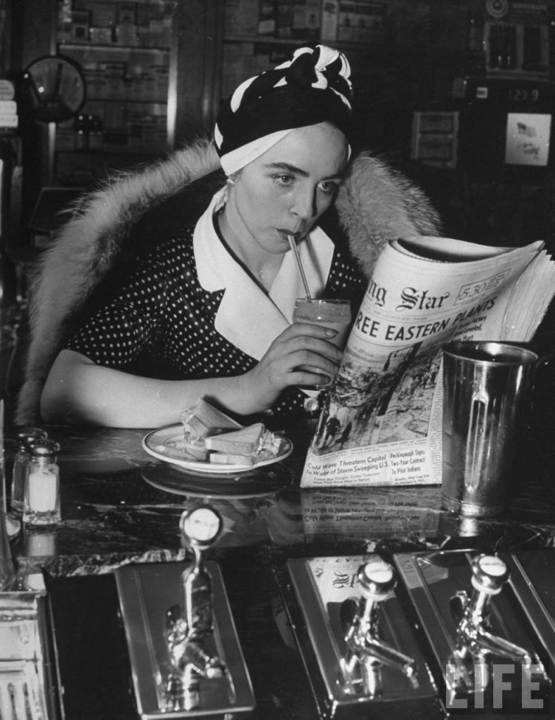 1940s vintage image of a woman wearing a fur coat and a 1940s style turban reading the newspaper at a malt shop counter. 