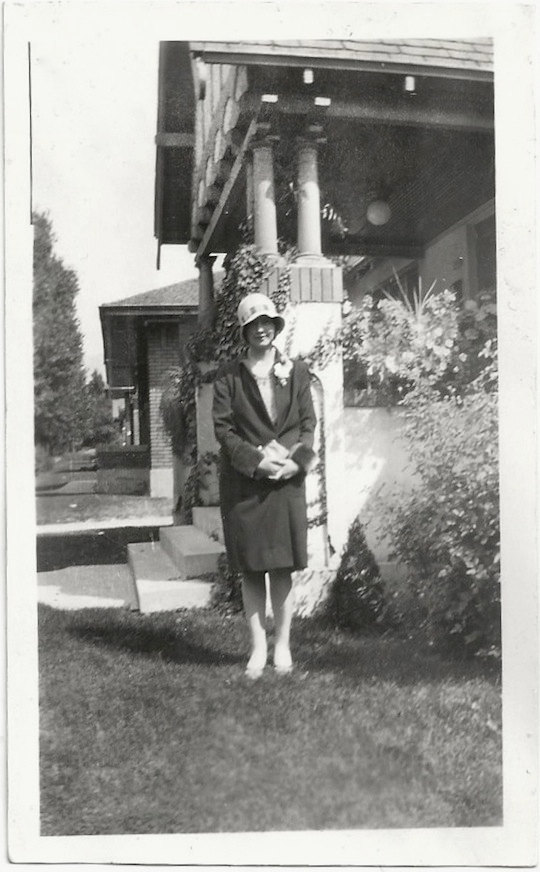 1920s vintage photo of a young woman in 1920s fashions (1920s coat, and 1920s hat) posing in front of her house. 