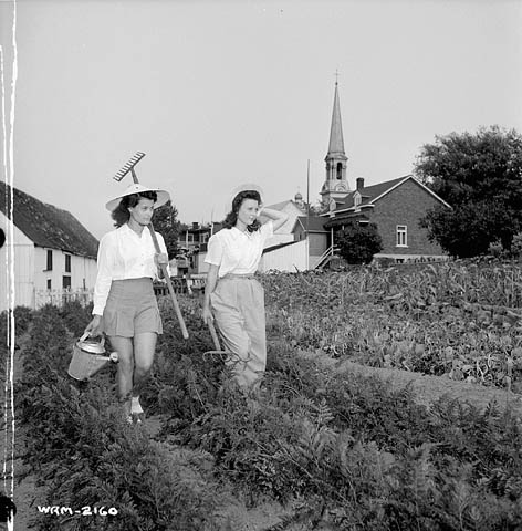 1940s vintage photo: ww2 canadian woman working for the war effort -The Perry Sisters, employed at the Dominion Arsenals Ltd. plant, armed with rake, watering can and pitchfork, help look after the vegetable garden where they are working