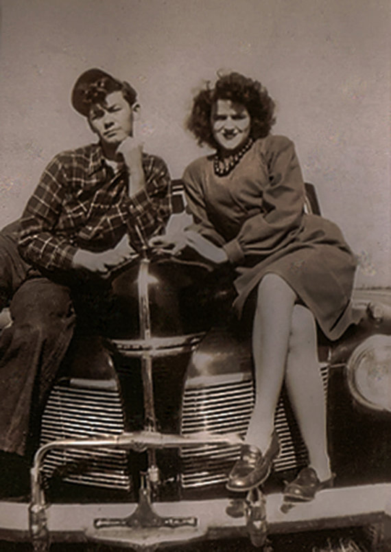 1940s vintage photo of a young couple in 1940s fashions posing on a car. 