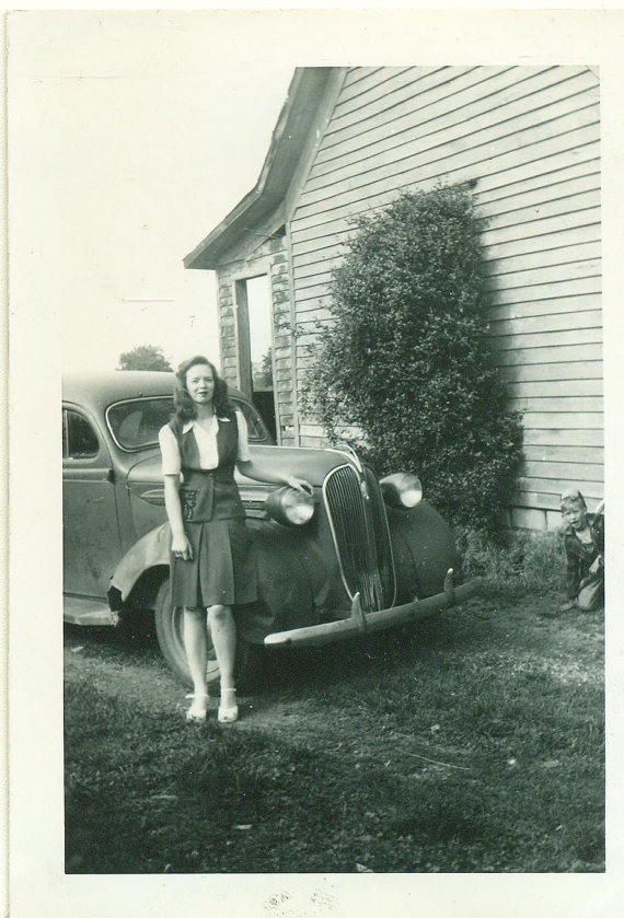 1940s vintage photo of a young woman in 1940s fashion posing in front of a car. 