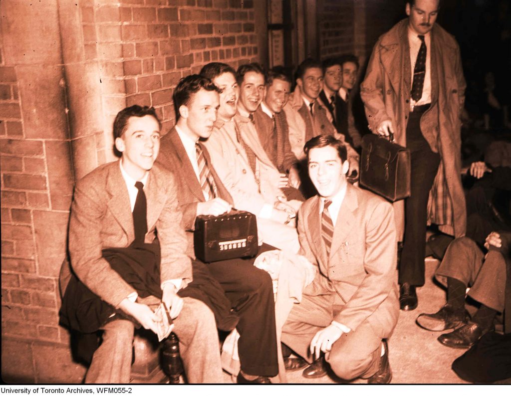 1940s vintage image of 1948 image of young men with a portable radio in suits - Boys and their portable radio-1948. Lineup outside the Athletic office for tickets for McGill football weekend.