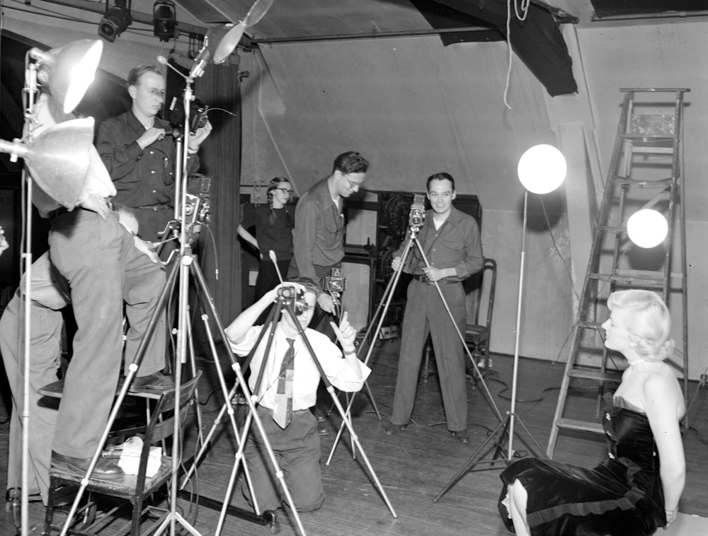 1950's photo of the Camera Club at the University of Chicago taking a picture of a pretty young woman in a stylish 1950s dress and hairstyle. 