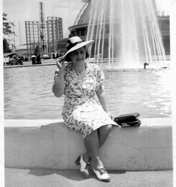 1940s vintage photo of a woman in a 1940s dress, wearing a hat and fantastic 1940s spectator style shoes posing in front of a water fountain. 