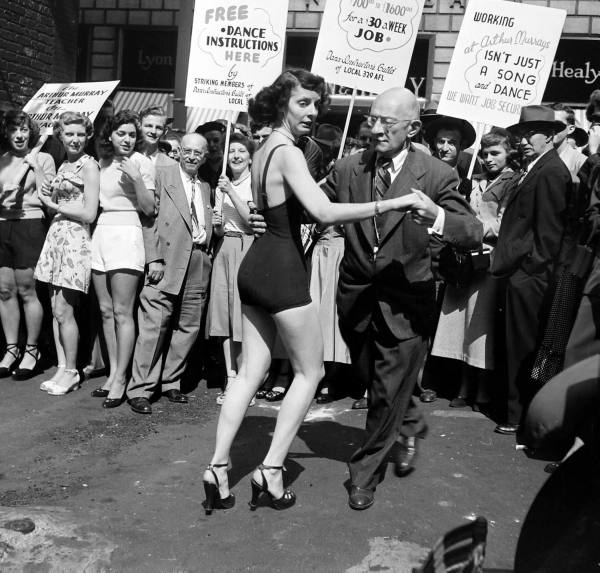 1940s Vintage Image of the Dancing Teachers Strike 1947. Image showcases strikers holding signs in the background while a woman in a swimsuit partner dances with a man.