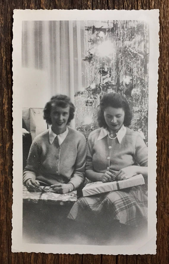 1940s vintage image of two women at christmas in cardigans and plaids skirts opening gifts in front of the tinsel Christmas Tree. 