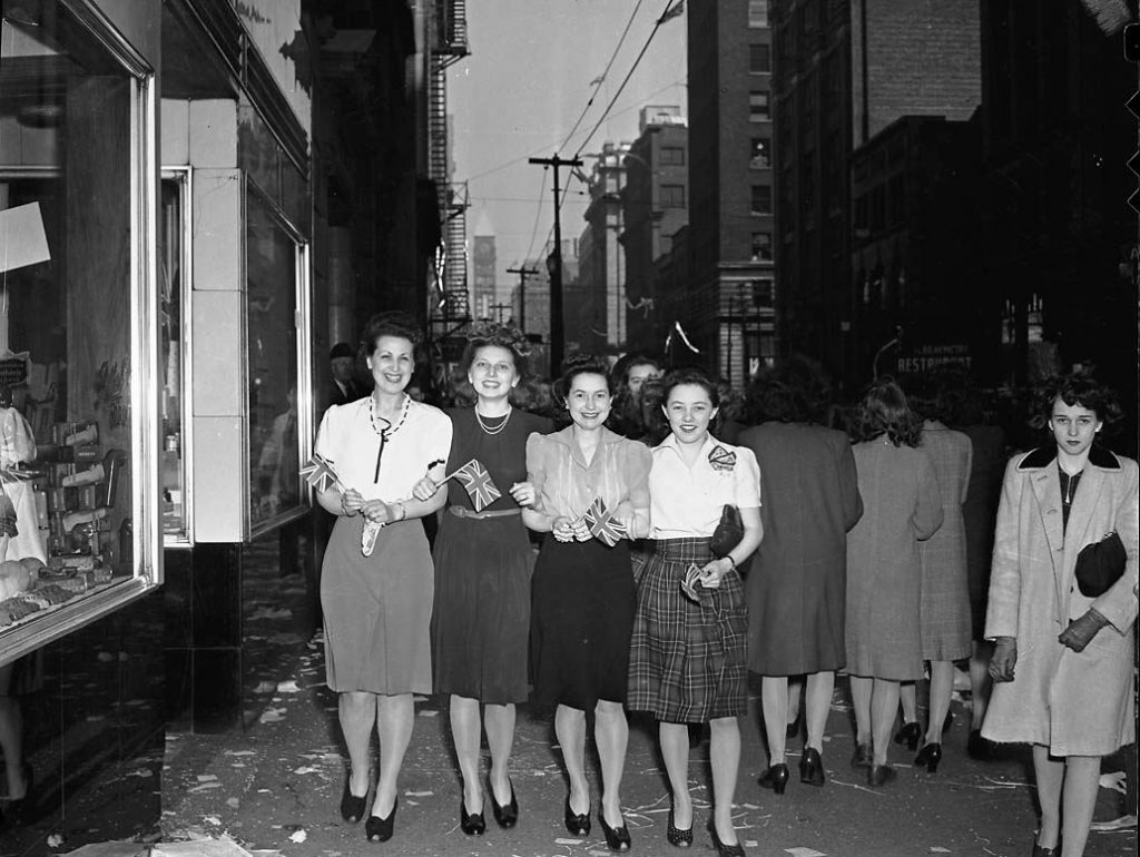 1940s vintage photo of Celebrating VE Day on Bay Street in Toronto. The image features 4 women hand in hand walking down the street in 1940s fashions. 