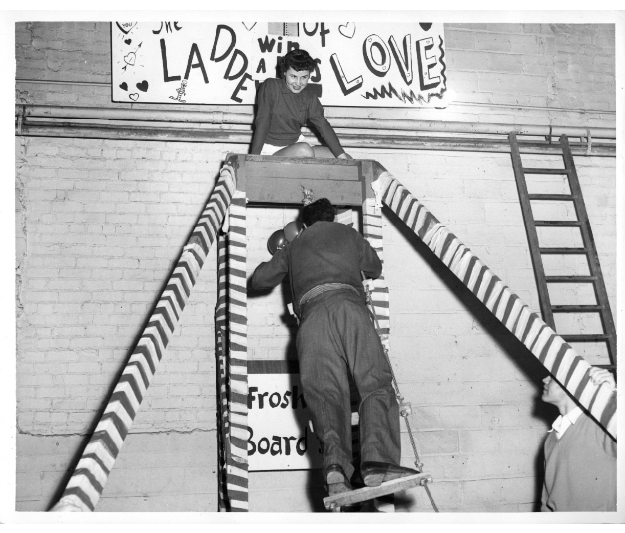 1950s vintage photo of student life, A Frosh Board game that features a man climbing a rope ladder to reach a woman. It advertises the prize of "Win a Kiss."