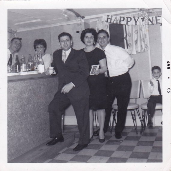 1960s vintage photo of a basement New Years Eve Party featuring a group standing around a 1960s home bar with a little kid sitting down in 1965. 