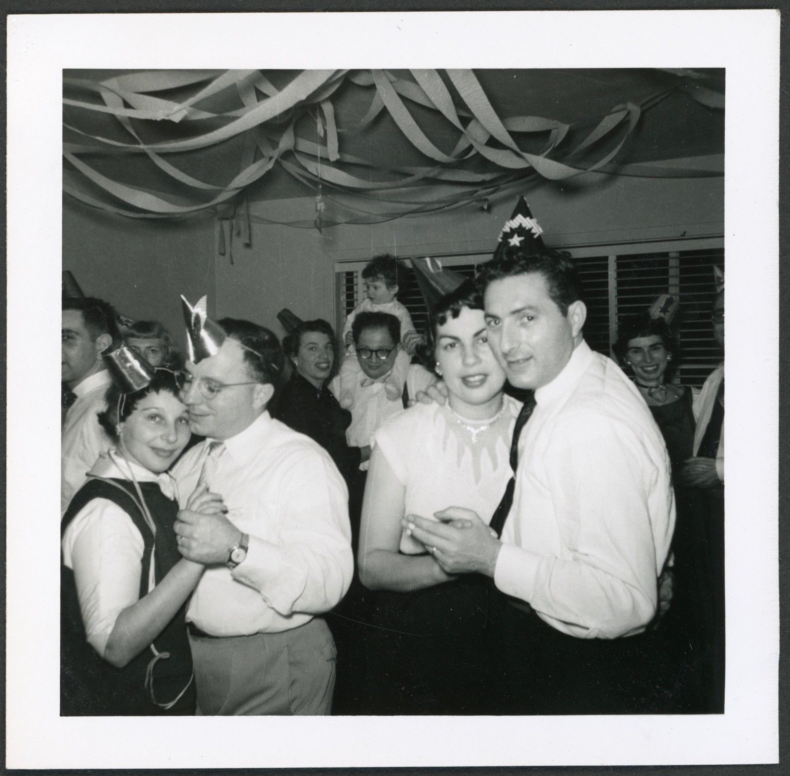 1950s Vintage photo from 1954 of a house party featuring a group of party goers in 1950s fashions and 1950s party hats dancing. 