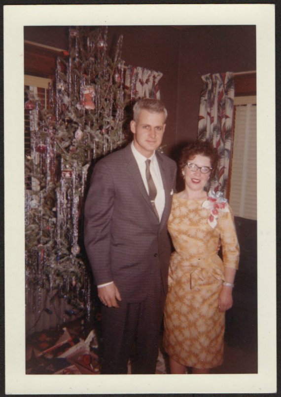 1960s vintage photo of a couple in 1960s fashions and a woman wearing a Christmas corsage posing by their Christmas Tree. 