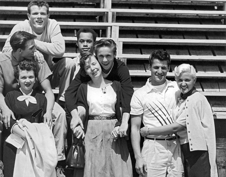 1950s vintage photo of a group of teenagers posing for a picture in the football stands. They are wearing typical 1950s fashions for teens at time. 