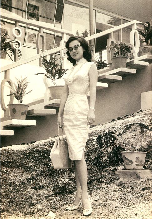 1950s vintage photo of a stylish 1950s Asian women in a 1950s traditional dress-Cheongsam with 1950s cat eye glasses and mid century architecture in the background. 