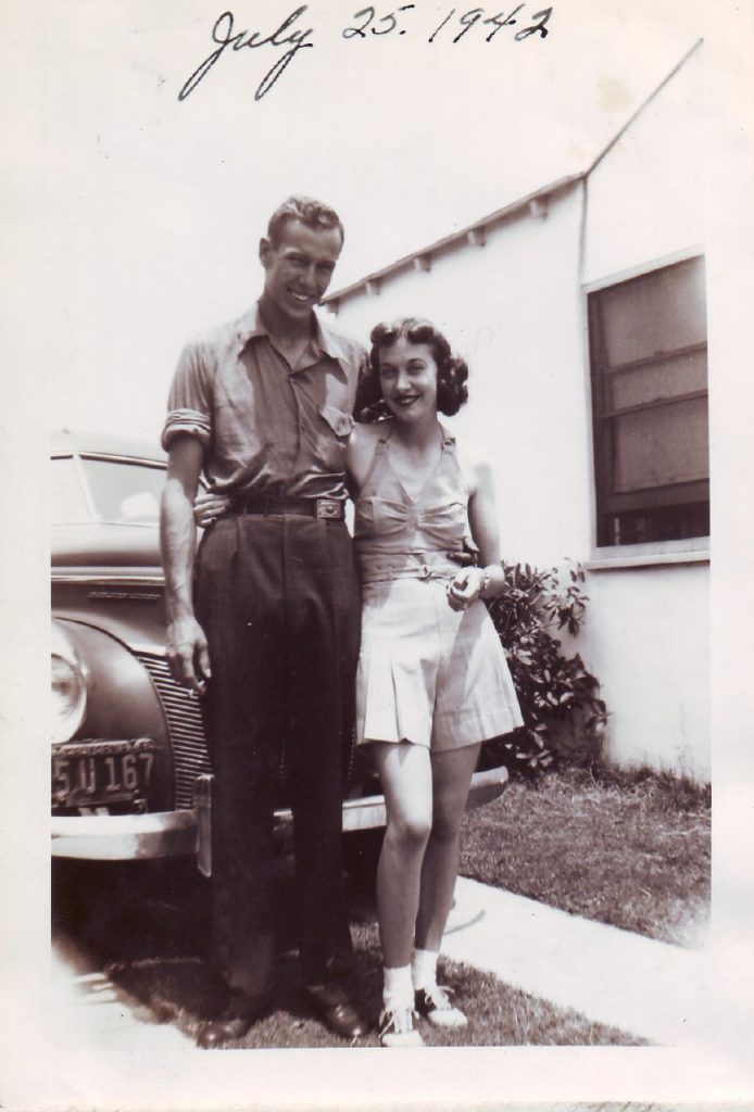 1940s Vintage photo of a young couple posing in front of their car in 1940s fashions / 1940s summer fashions. The young woman is wearing victory rolls and a playsuit and saddle shoes. 