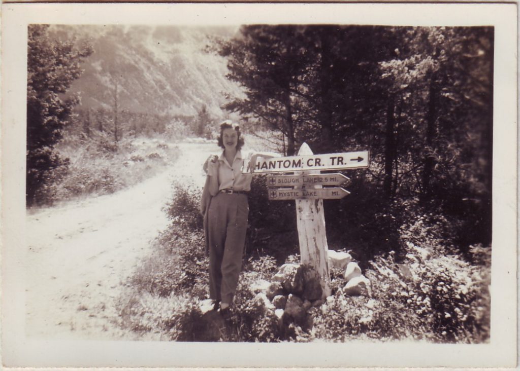 1940s vintage photo of a young woman on a trip posing by a sign in 1940s fashion (1940s pants and blouse and 1940s hairstyle). 