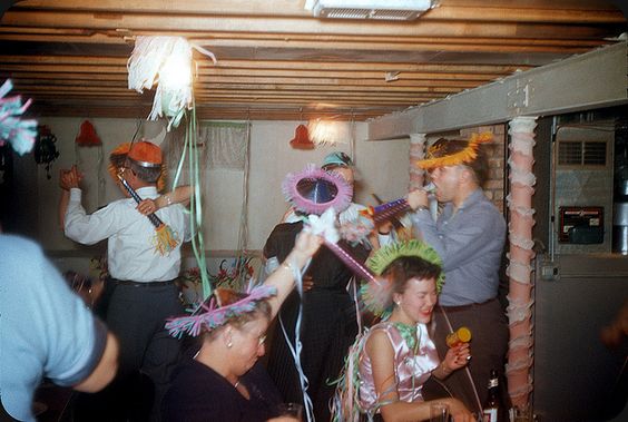 1950s vintage photo of a New Years Party in the basement of a house featuring a group of party goers in party hats and 1950s fashions dancing and having fun. 