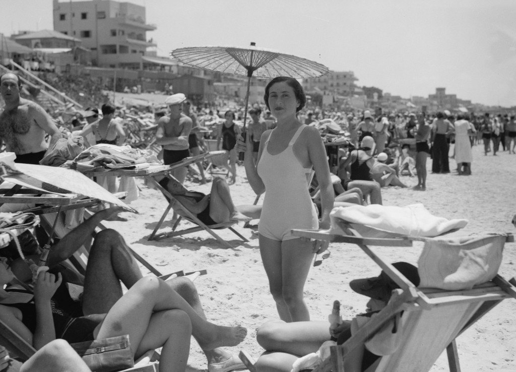 1930s vintage photo: Woman on beach in Tel Aviv in 1934. She is holding a parasol while posing on a busy beach in her outstanding 1930s bathingsuit. 