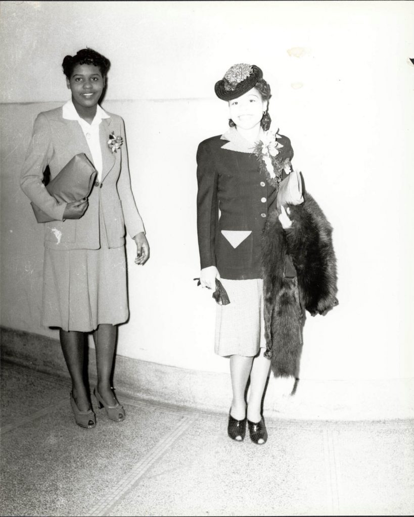 Two Black women, including Velma Pratt Bell, holding handbag and fur, standing in Schenley High School for Beauty Shop Owners' Fashion Revue, circa 1945. Photo by Teenie Harris. They are wearing stylish Skirt Suits