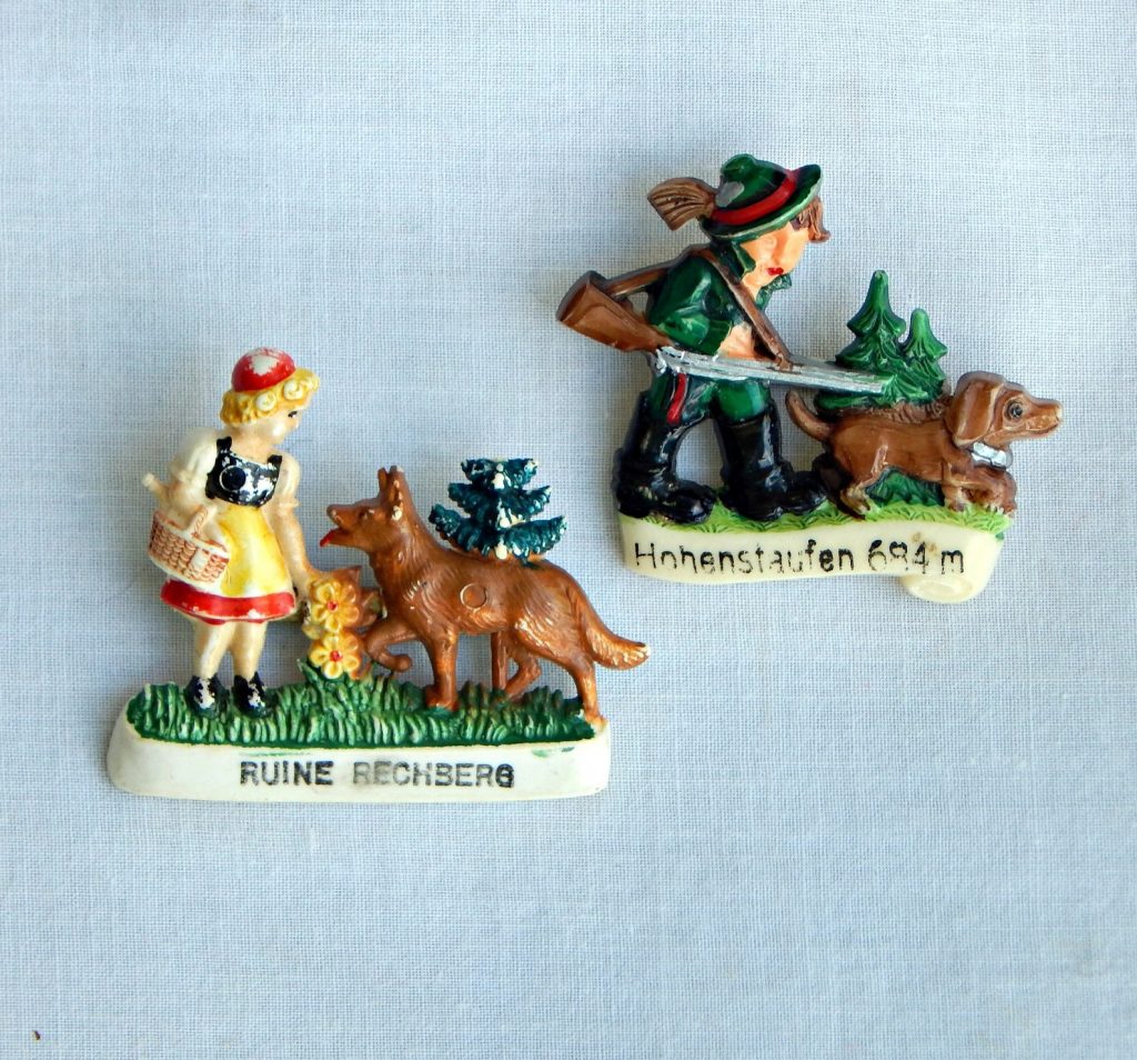 1950s Hat Pins: The pin from Hohenstaufen has an alpine hunter with his dog. The pin from Rune Rechberg has a girl in tradional dress holding flowers with her dog nearby.