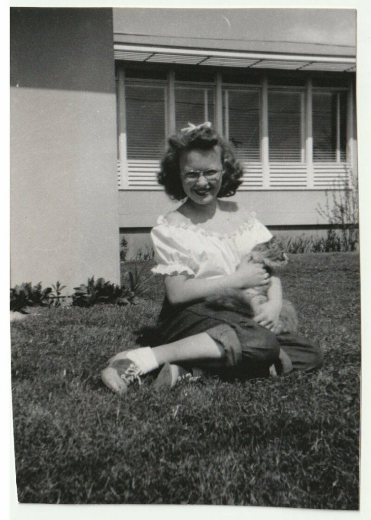 1940s photo of a young girl on the front lawn in saddle shoes, glasses and 1940s vintage hairstyle with her cat. 