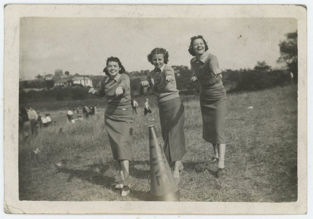 Early 1940s Photo of 3 women in long pencil skirts and letterman sweaters getting their cheer on! The woman on the left is wearing saddle shoes.