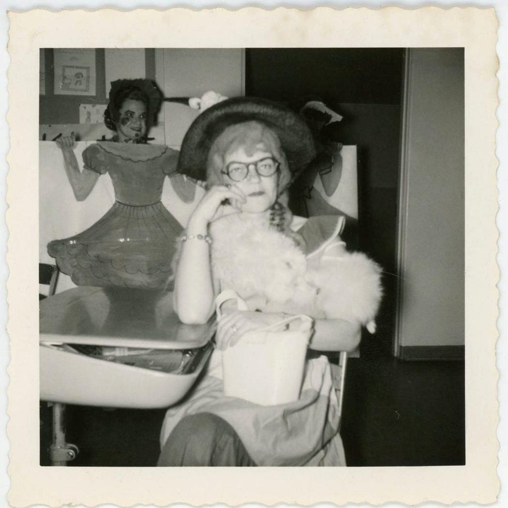 1960s photo of a school setting Halloween party~one woman has her head above an arcade type cut away in back. Lady at desk isn't sure who she is with her fur stole.