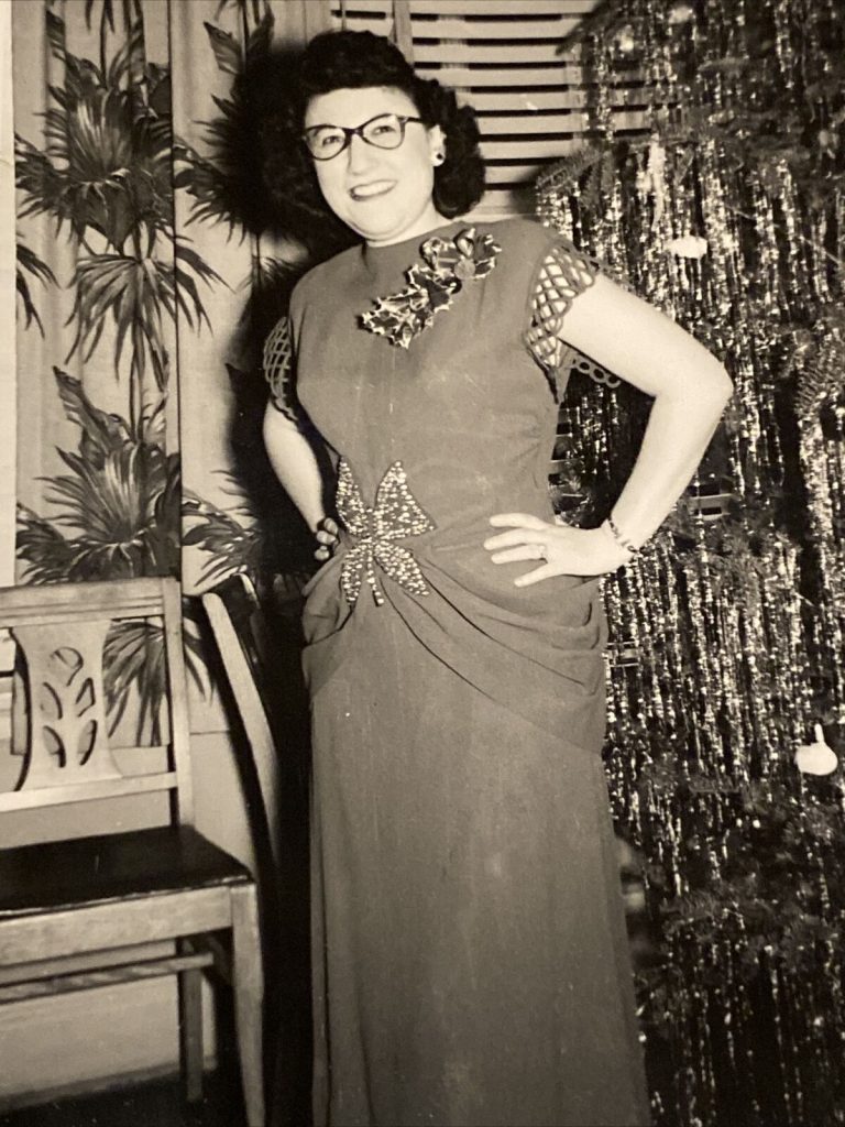 1940s vintage photo of a women in a 1940s evening gown with large christmas brooch wearing glasses and standing in front of a tinsel tree