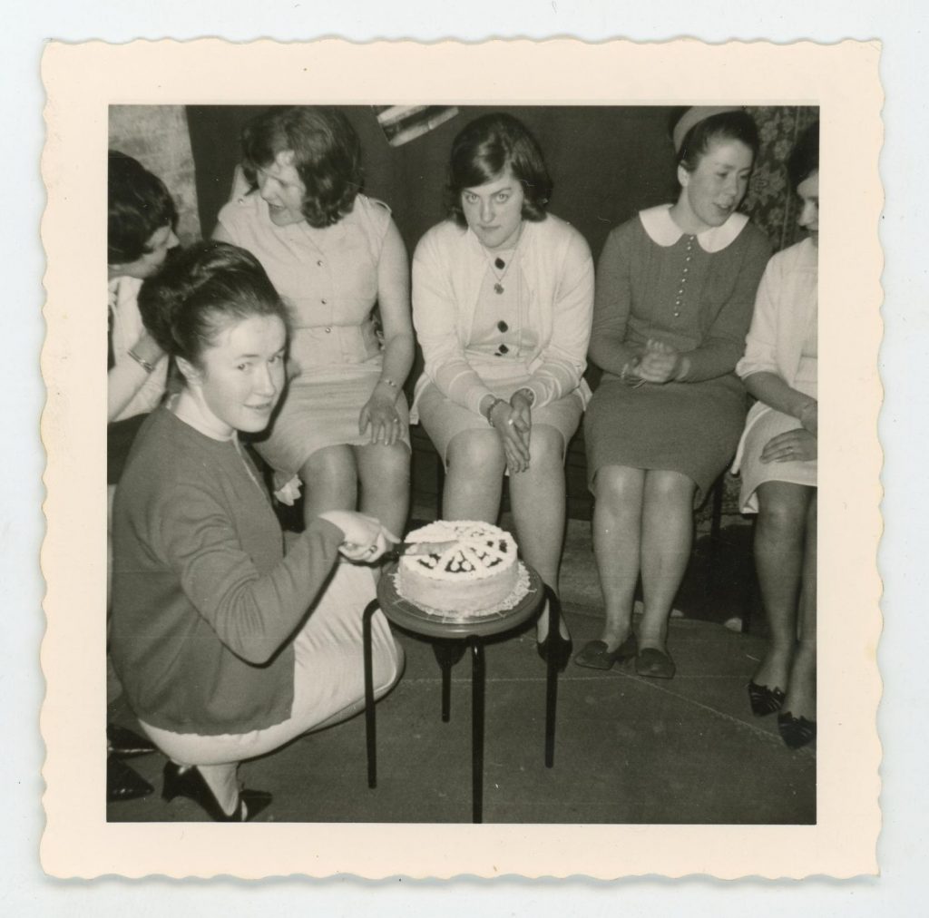 1960s vintage photo of a woman cutting a cake with a group of other women watching on in 1960s fashions