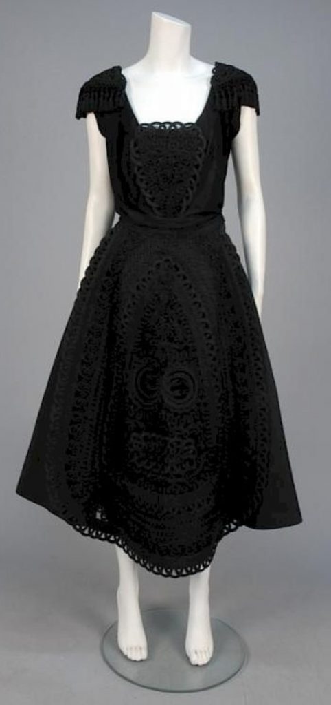 1950s Fashion: 1953 Cocktail Dress. Black jersey sleeveless bodice with bib of black braid and cord, faille circle skirt, braid and cord decorated net apron