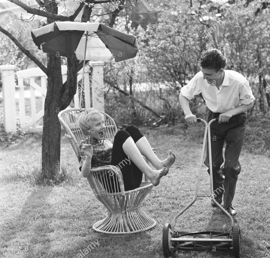 Summer activitiy in the 1950s. A young woman sitting in typical 1950s garden chair holds her feet up so her partner can cut the grass just where she is sitting. Sweden May 1959