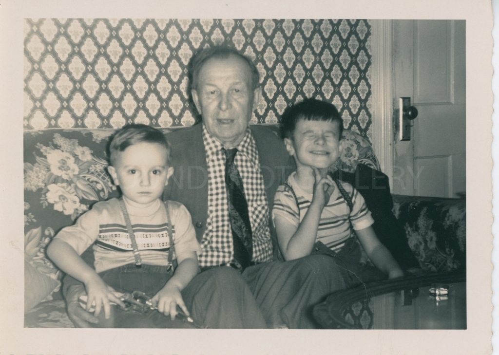 1950s vintage photo of a 2 young boys with their grandfather sitting on the couch in front of a wallpaper wall. 