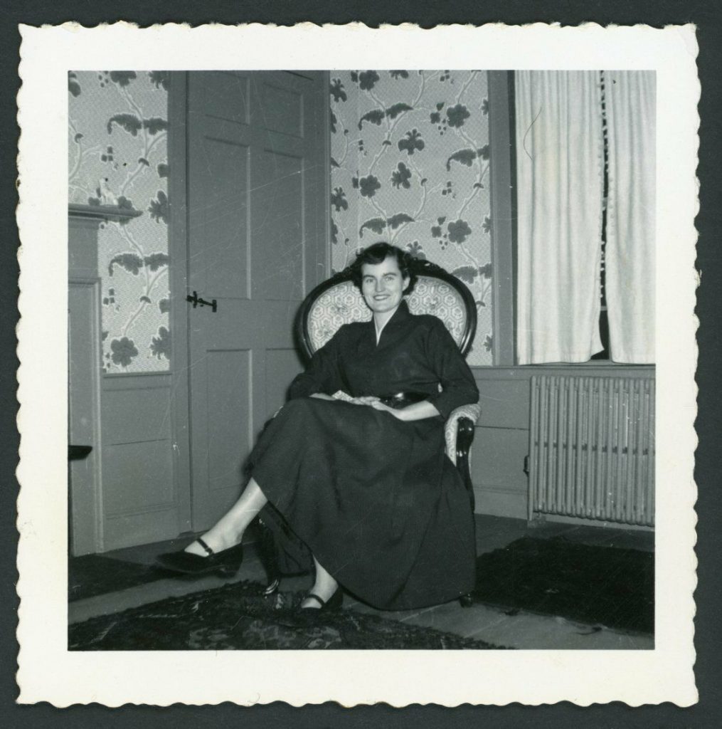 1950s vintage photo of a woman in a 1950s dress sitting in chair in front of a wall with wallpaper. 