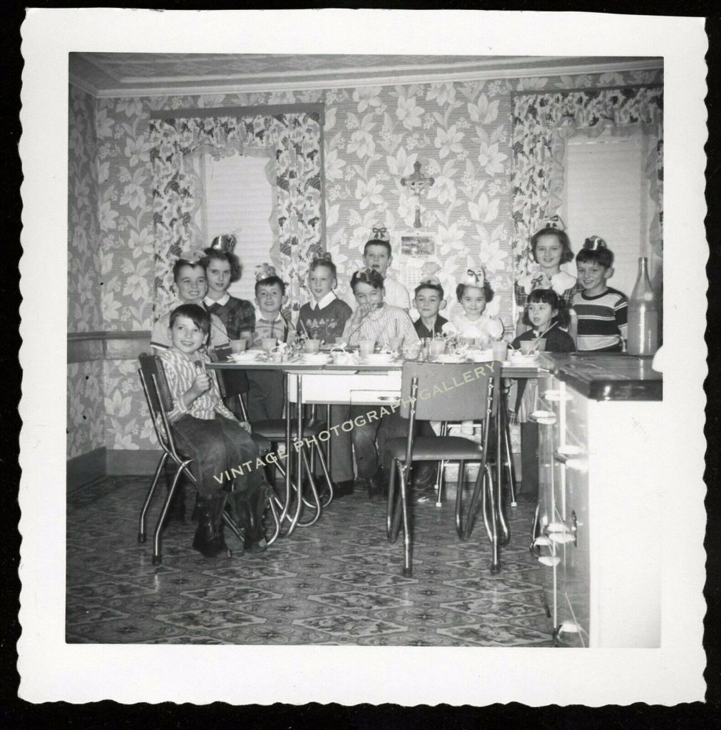 1950s / 1960s vintage photo of a kids birthday party in a room with wallpaper and floral curtains plus a formica table. 