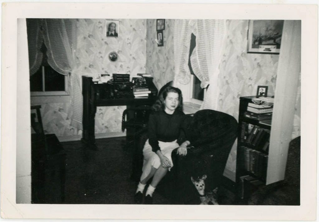 1950s vintage photo of a young girl sitting in a chair in a livingroom with a toy deer at her foot and a wall covered in wallpaper. 