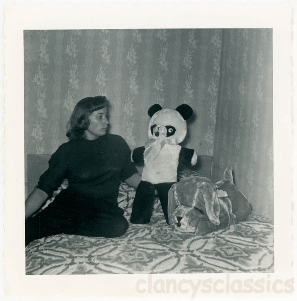 1950s vintage photo from 1955 of a young woman in a bedroom with a stuffed panda bear and stuff dog sitting in front of a room with wallpaper. 