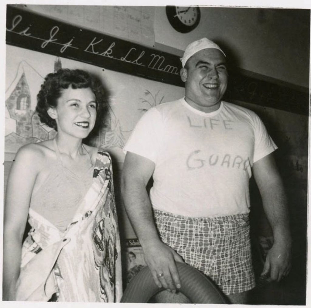 From a Halloween costume party at a school, a man in a lifeguard costume and woman in her swimsuit in this 1950s vintage photo