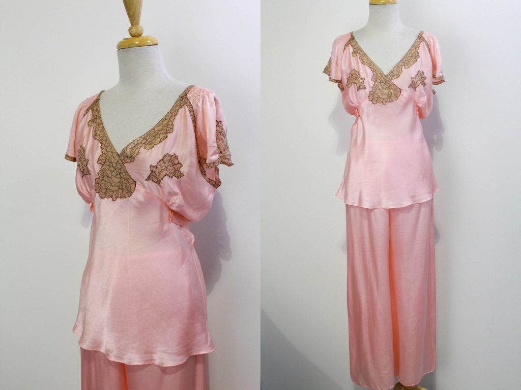 1940s Fashion: Beautiful pink silk satin pyjama set by Dunning Lingerie from the 1940s. The top has lace detailing on the neckline and bust and an empire waist that ties in the back. The neckline dips into a V in the front and back, and the fluttery sleeves are detailed and trimmed with lace. The pants have sweeping wide legs and a slight elasticized waist.