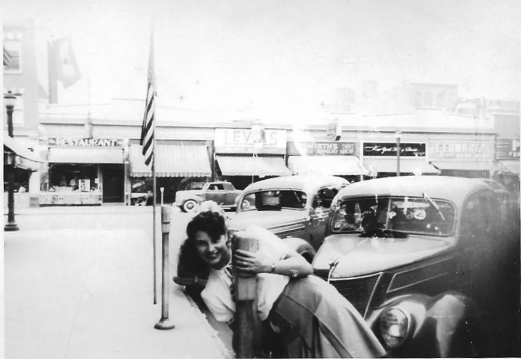 1940s Vintage Photo of a woman posing in front of cars parked on the street in main street shopping area. 