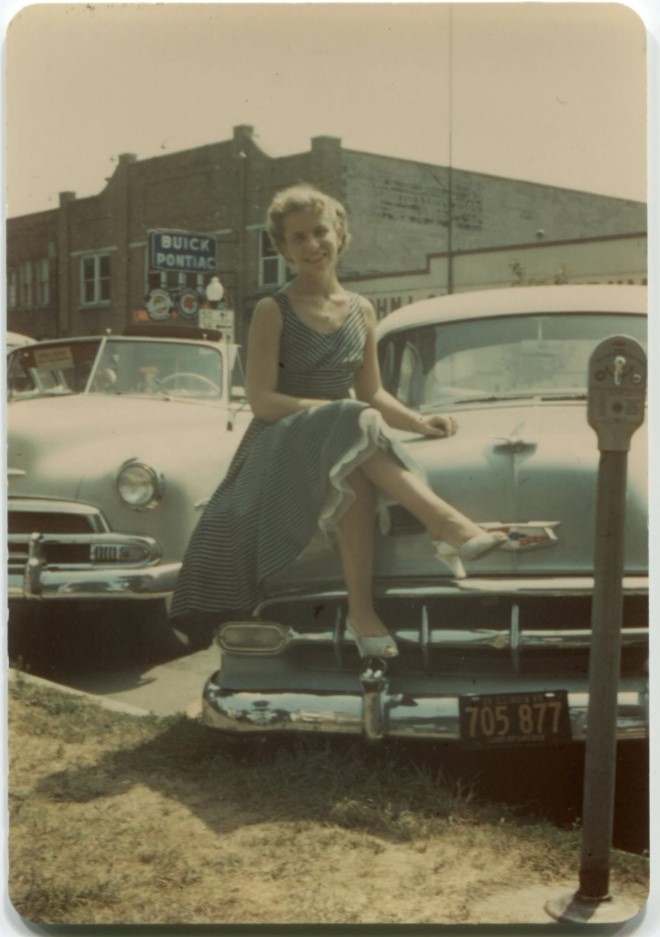 1950s colour photo of a women in 1950s dress sitting on the hood of a car