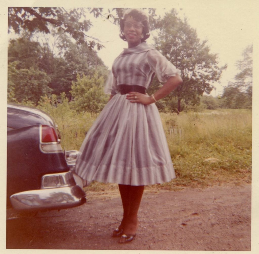 1960s vintage photo of a Black Women in a stylish early 1960s dress and cat eye glasses posing behind a car. 